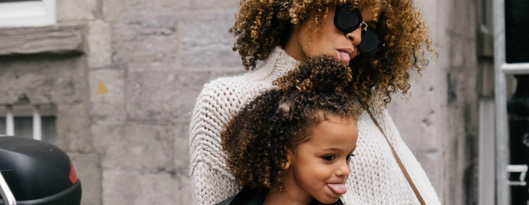 mother and daughter stick their tongues out while looking at themselves in a motorcycle mirror parked in front of a building