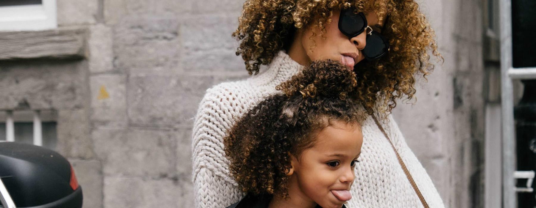 mother and daughter stick their tongues out while looking at themselves in a motorcycle mirror parked in front of a building